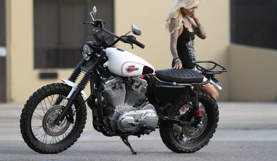 A woman in a black dress is standing next to a motorcycle at home.