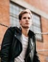 A young man in a leather jacket leaning against a chain link fence, giving a testimonial.